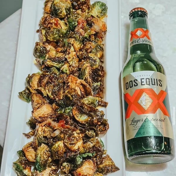 Crispy Brussels sprouts with bacon on a white platter next to a bottle of Dos Equis Lager Especial.