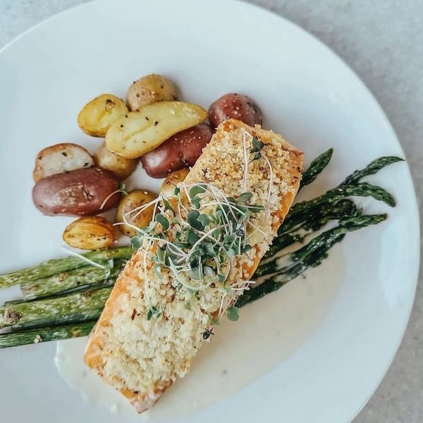 Breaded salmon fillet topped with microgreens, roasted potatoes, and grilled asparagus on a white plate.