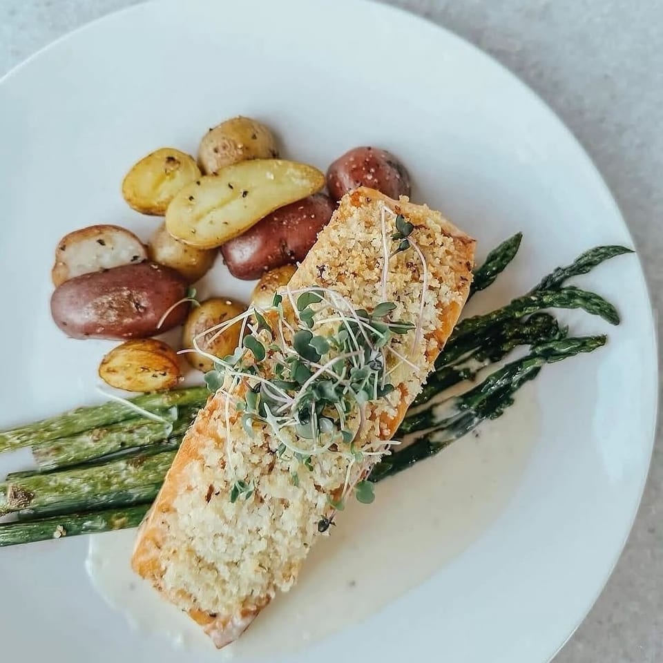 Breaded salmon fillet topped with microgreens, roasted potatoes, and grilled asparagus on a white plate.