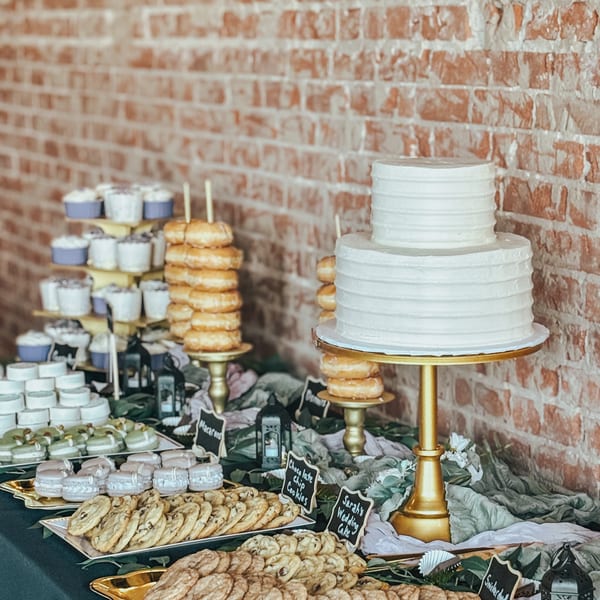Dessert table featuring a white tiered cake, stacked doughnuts, cupcakes, cookies, and labeled treats against a brick wall.