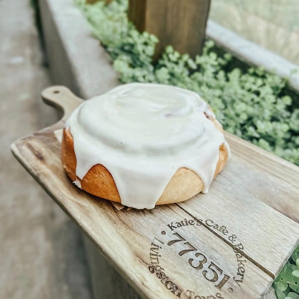 Round golden cake drizzled with white icing on a wooden Katie’s Café and Bakery serving board, surrounded by greenery.