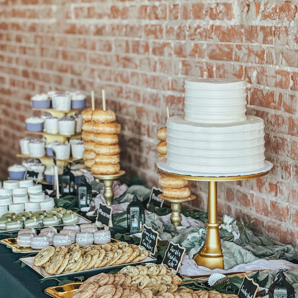 A dessert table with a two-tiered white cake, donuts, macarons, cupcakes, and cookies.