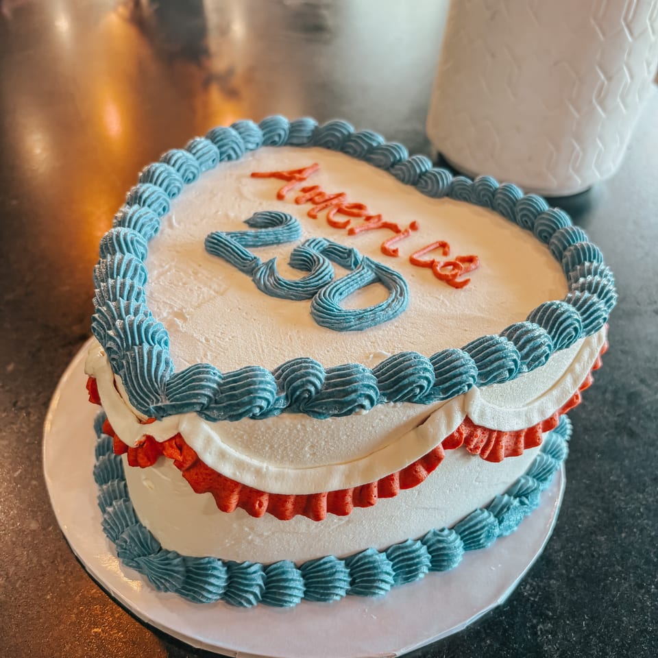 Heart-shaped cake with white frosting, blue and red trim, and "America 2023" written on top.