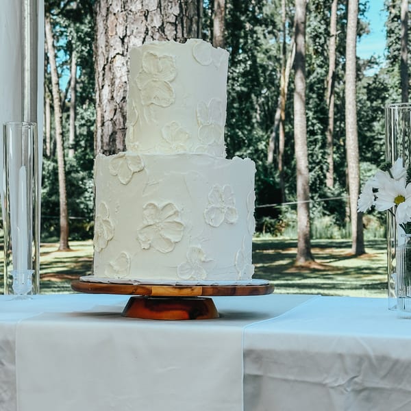 A two-tiered white wedding cake with floral icing sits on a wooden stand by a window overlooking trees.