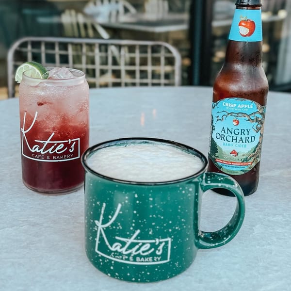 Katie's Cafe & Bakery branded drinks: a red iced beverage, a foamy beer mug, and an Angry Orchard cider.