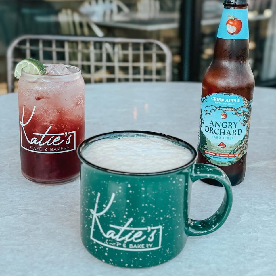 Katie's Cafe & Bakery branded drinks: a red iced beverage, a foamy beer mug, and an Angry Orchard cider.