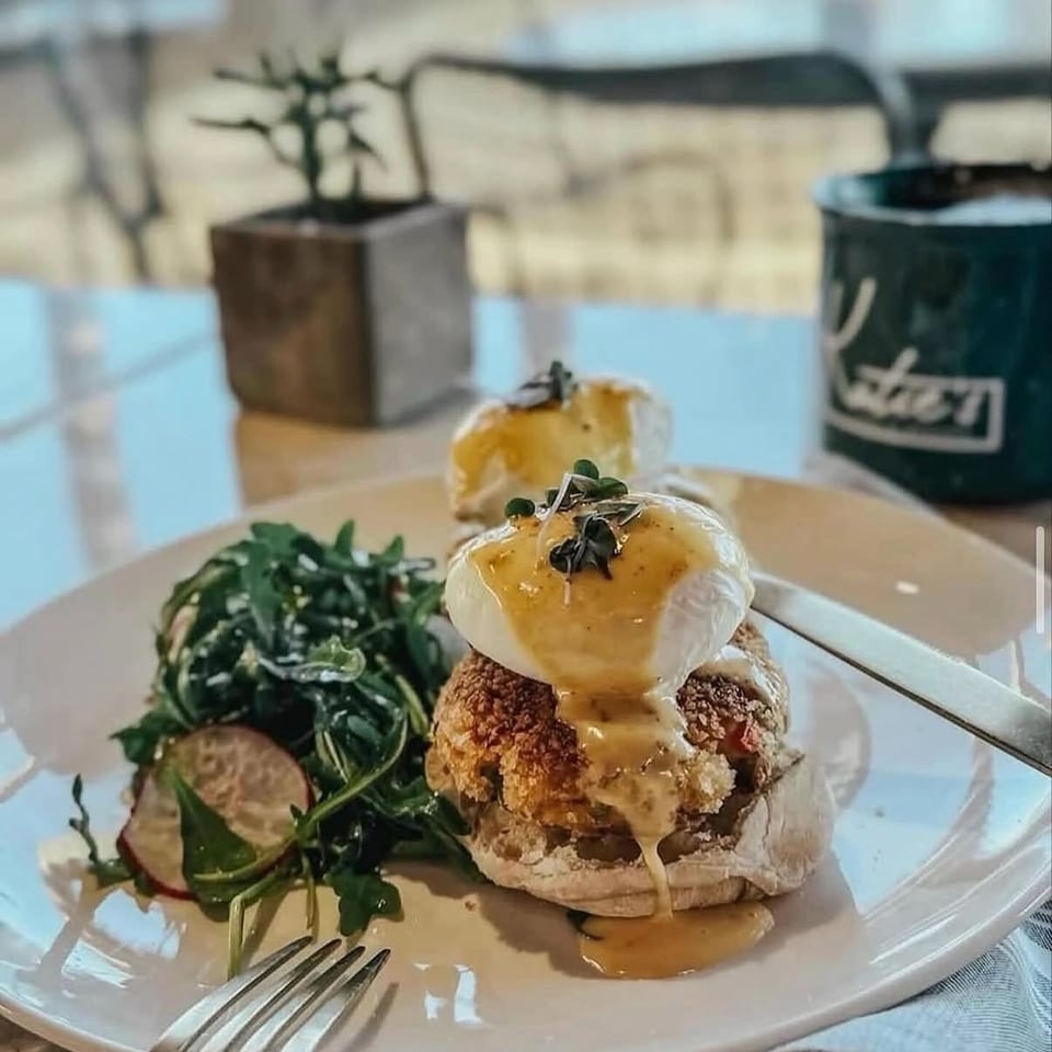 Crispy breaded sandwich topped with poached eggs and creamy sauce, served with greens and sliced vegetables on a plate.